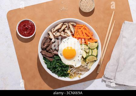 Vista dall'alto di una ciotola bibimbap coreana con manzo, verdure e uova fritte, scattata con una luminosa illuminazione da studio su uno sfondo neutro Foto Stock