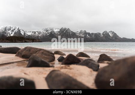 Ambientazione invernale su una spiaggia sabbiosa nelle isole Lofoten, Norvegia, con montagne innevate sullo sfondo. Il primo piano Rocky aggiunge profondità al tr Foto Stock