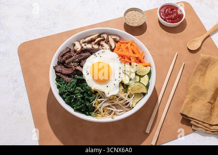 Vista dall'alto di una ciotola bibimbap coreana con manzo, verdure e uova fritte, scattata con una luminosa illuminazione da studio su uno sfondo neutro Foto Stock