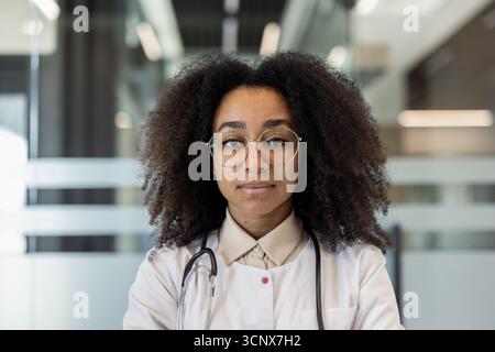 Giovane medico donna che indossa occhiali e stetoscopio, trasudando fiducia e professionalità. Racchiuso in un moderno ambiente di studio medico, rappresenta la diversità e la competenza nel settore sanitario Foto Stock
