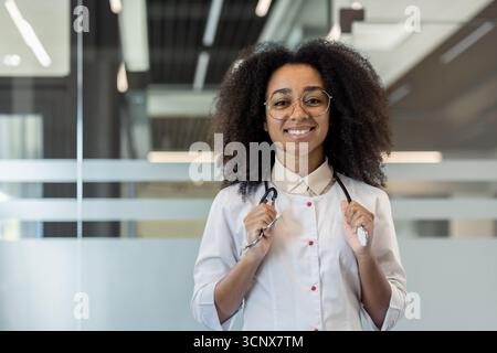 Ritratto di una giovane donna afroamericana sorridente in piedi in una hall dell'ospedale, tenendo uno stetoscopio e guardando la macchina fotografica. Foto Stock