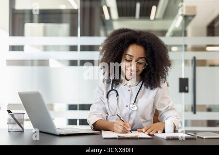 Una giovane dottoressa afroamericana è seduta a una scrivania in un abito e lavora su un computer portatile e con i documenti. Foto Stock