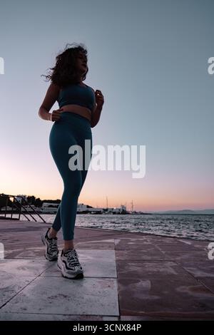 Una giovane donna con i capelli bruna corre lungo il mare al tramonto, indossando un abbigliamento sportivo blu. Il mare tranquillo e il cielo colorato creano un'atmosfera stimolante Foto Stock