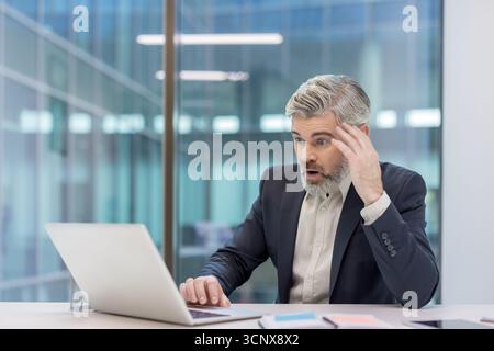Uomo d'affari maturo che soffre di shock e incredulità guardando lo schermo di un computer portatile, reagendo a cattive notizie inaspettate o a un evento sorprendente in un ufficio moderno Foto Stock