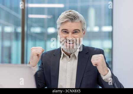 Uomo d'affari anziano con i capelli grigi e un sorriso che solleva i pugni stringuti, esprimendo entusiasmo e risultati in un moderno ambiente d'ufficio Foto Stock