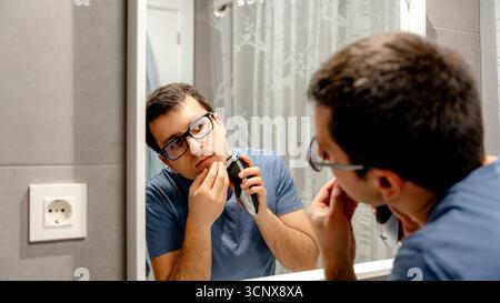 Un uomo con una camicia blu si rasa la barba con un rasoio elettrico mentre guarda il suo riflesso nello specchio del bagno. La scena cattura una routine gr Foto Stock
