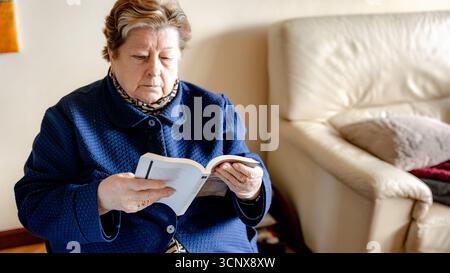 Una donna anziana legge un libro mentre è seduta al chiuso, indossando una sciarpa e una giacca calda. La scena evoca tranquillità, concentrazione e divertimento Foto Stock
