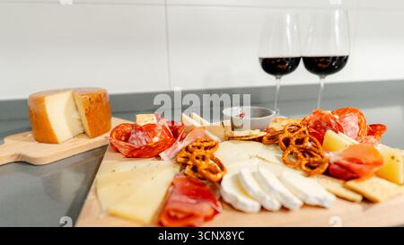 Un'ampia scelta di formaggi assortiti, affettati, cracker e pretzel preparati ad arte su una tavola di legno, accompagnati da due bicchieri di vino rosso, Foto Stock