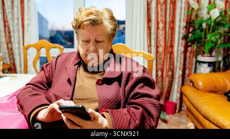 Una donna anziana seduto comodamente a casa, concentrandosi sul suo smartphone. L'ambiente e' intimo e caldo, con tende colorate e decor che creano un benvenuto Foto Stock