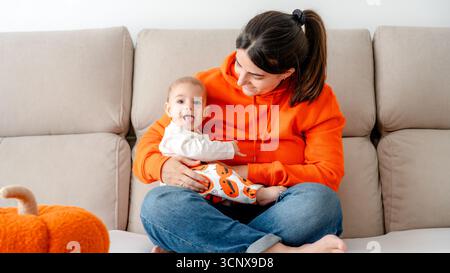 Una madre gioiosa e il suo bambino di 4 mesi, in abito festoso di Halloween, godetevi momenti divertenti sul divano in un accogliente soggiorno con connessione wi-fi Foto Stock