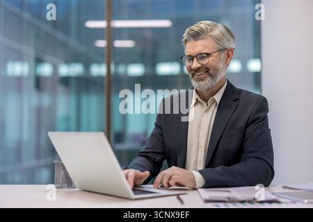 Uomo d'affari anziano sorridente, in rete e che lavora con un laptop a una scrivania, circondato da documenti d'ufficio e che riflette l'architettura moderna Foto Stock
