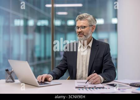 Uomo d'affari professionista esperto che lavora alla scrivania, digita sul computer portatile e rivede grafici e report in un moderno ambiente aziendale Foto Stock