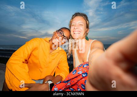 Due giovani donne sorridenti, una caucasica e una nera, si fanno un gioioso selfie su una spiaggia soleggiata. Catturano un momento felice di amicizia contro un bea Foto Stock
