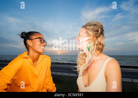 Due giovani e allegri donne multietniche si godono un momento vibrante in riva al mare. Una donna nera e una donna caucasica condividono una risa gioiosa, legandosi nel W Foto Stock