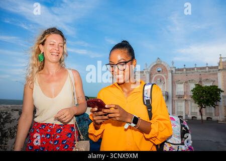 Due giovani donne multietniche esplorano la splendida posizione all'aperto. La donna nera usa uno smartphone mentre la donna caucasica ride, condividendo una gioiosa m Foto Stock