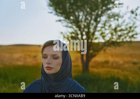 Una donna in un hijab blu si trova in un tranquillo prato estivo. La luce soffusa e dorata del sole bagna il paesaggio, con un albero solitario sullo sfondo che completa il Foto Stock