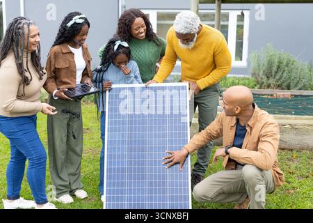 Famiglia diversificata con tecnici solari che esaminano pannelli solari di grandi dimensioni e piccoli moduli su prato erboso Foto Stock