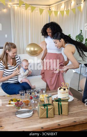 Diverse amiche con neonati che festeggiano il 30° compleanno in soggiorno, torta e regali Foto Stock