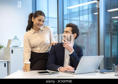 Una felice coppia d'affari che lavora insieme in un ufficio aziendale, condividendo un momento di connessione spensierata con sorrisi, dimostrando dinamiche di lavoro positive Foto Stock