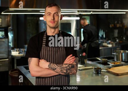 Ritratto di un giovane cuoco caucasico con tatuaggi in piedi in cucina professionale che incrocia le braccia e sorride con fiducia mentre un altro chef lavora in background Foto Stock