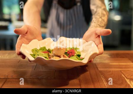 Giovane uomo caucasico con braccia tatuate che presenta un piatto gourmet con verdure a foglia verde e carne su piatto decorativo, in piedi dietro il bancone in legno nella cucina del ristorante Foto Stock