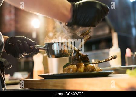 Giovane uomo caucasico adulto che indossa guanti neri che ricopre costolette di agnello e verdure cotte nella cucina del ristorante, tenendo in mano la pentola e il cucchiaio mentre si aggiunge la salsa al piatto, il vapore che fuoriesce dal cibo Foto Stock