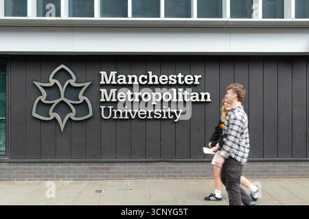 Due giovani studenti adulti che oltrepassano il cartello della Manchester Metropolitan University su una facciata moderna dell'edificio MMU Foto Stock