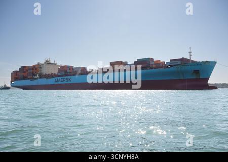 Una grande nave container di Maersk che trasporta numerosi container Hapag-Lloyd e Maersk naviga attraverso il mare, accompagnata da un piccolo rimorchiatore. Foto Stock