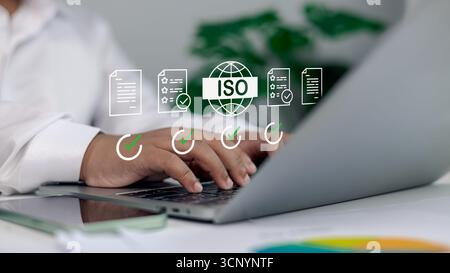 Primo piano di un uomo d'affari che scrive su un laptop con icone di certificazione ISO, lista di controllo dei documenti e segno di spunta verde, concetto di gestione della qualità, standard Foto Stock