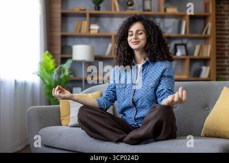 Donna a occhi chiusi e espressione serena che gode di un momento di consapevolezza e armonia mentale, pratica yoga e tecniche di rilassamento nel suo salotto tranquillo Foto Stock