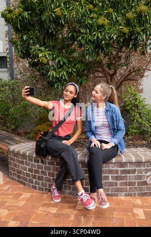 Diverse amiche che posano sulla panchina di piantagione del cortile con telefono e borsa che scattano selfie Foto Stock