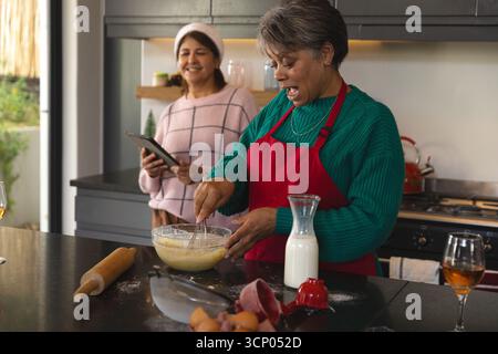 Mamma e figlia che sbattono l'impasto nel recipiente e controllano la ricetta sul tablet all'isola della cucina Foto Stock