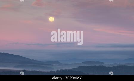 La luna piena si illumina sopra un paesaggio nebbioso, emettendo una luce morbida ed eterea sulla scena. Foto Stock