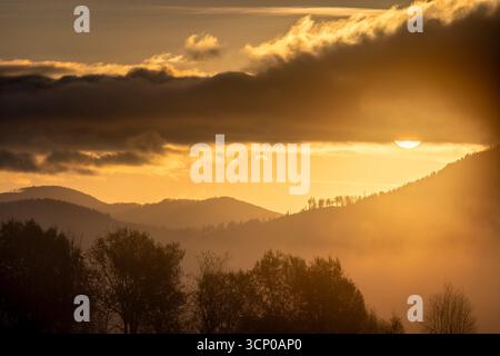 L'ora d'oro bagna il paesaggio montuoso con un bagliore nebbioso mentre il sole si infrangeva tra le nuvole. Foto Stock