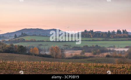 Colline ondulate e campi autunnali sotto un cielo pastello, creando un tranquillo paesaggio rurale. Foto Stock