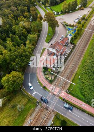 Vista aerea di un passaggio ferroviario diagonale in un'area semi-rurale, con auto fermate alle barriere, un edificio con tetto rosso con posti a sedere all'aperto, campo verde Foto Stock
