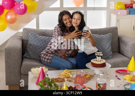 Mamma e figlia adolescente diverse si fanno selfie a casa con lo smartphone e la torta di compleanno Foto Stock
