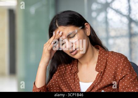 Giovane donna indiana con occhiali da vista e camicia a pois, che stringe la fronte in un ufficio, sopraffatta dal dolore simile all'emicrania, dallo stress e dalla stanchezza dovuti al lavoro eccessivo Foto Stock