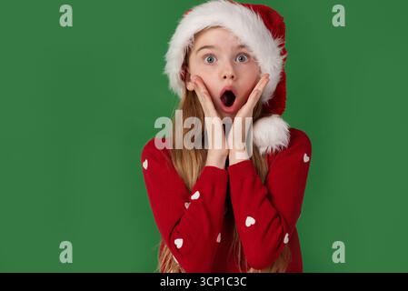 Una ragazza sorpresa con un cappello di Babbo Natale e un maglione rosso tiene la faccia sotto shock su uno sfondo verde Foto Stock
