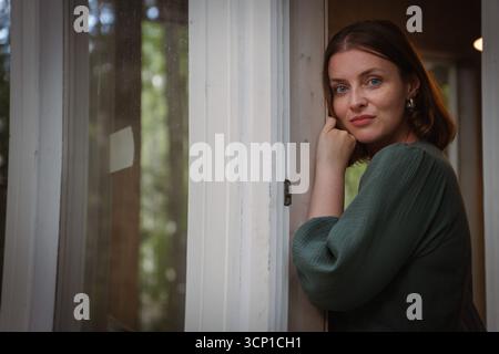 Una donna con un abito verde si appoggia a una porta, fissando delicatamente la fotocamera con un'espressione naturale e una luce calda Foto Stock