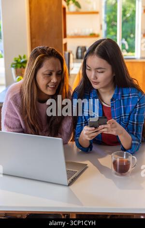 Madre e figlia asiatiche condividono lo smartphone mentre lavorano al laptop a tavola Foto Stock