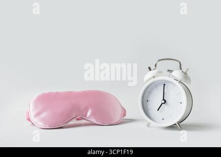 Maschera per gli occhi morbida e sagomata per un sonno confortevole. Maschera rosa e sveglia bianca su sfondo grigio. Concetto di sonno, insonnia, assistenza sanitaria, disordine quotidiano Foto Stock
