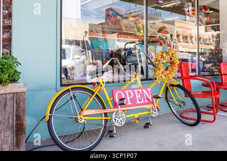 Lincolnton, Carolina del Nord, Stati Uniti - 14 luglio 2025: Bicicletta gialla costruita per due persone si appoggia contro il punto vendita, con un cartello "aperto". Foto Stock