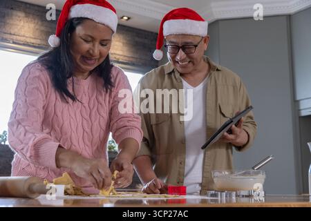 Diverse coppie anziane nei cappelli di babbo natale preparano l'impasto al piano di lavoro della cucina con un tablet Foto Stock