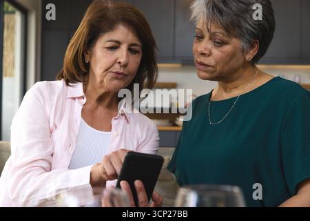 Diverse amiche anziane che puntano sullo schermo dello smartphone mentre sono sedute nella cucina di casa Foto Stock