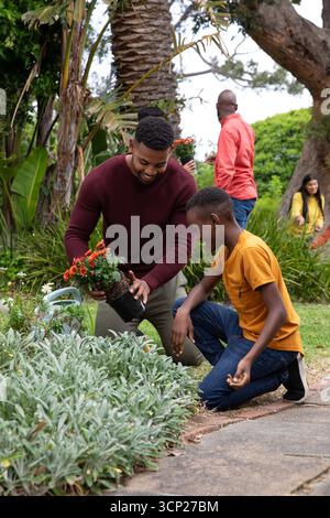 Padre e figlio afroamericani inginocchiati piantando fiori in vaso in cortile annaffiando lattine Foto Stock