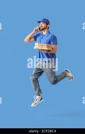Corriere maschile che salta con cassette per pizza che parla tramite telefono cellulare su sfondo blu Foto Stock