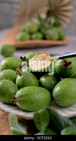 Uno shot ravvicinato ad angolo alto di una pila di frutti feijoa appena raccolti, alcuni con scintillanti goccioline d'acqua, su un piatto bianco. Foto Stock