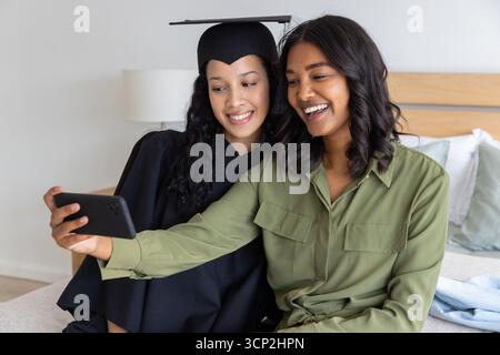 Mamma e figlia adolescente diversificate che scattano selfie in camera da letto con cappello, abito e smartphone Foto Stock