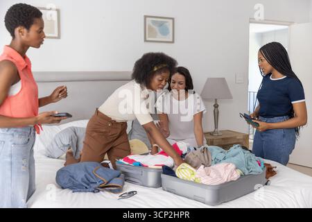 Diverse amiche che confezionano vestiti in valigie aperte sul letto con smartphone e tablet Foto Stock